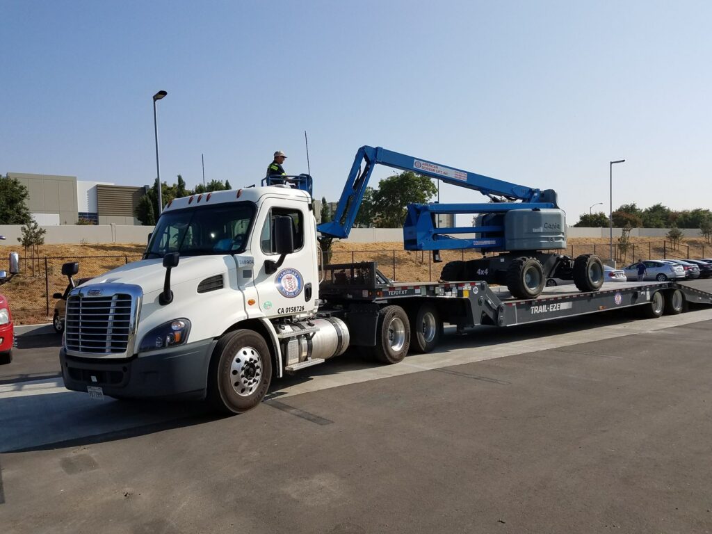 American Scissor Lift truck hauling a blue Genie articulating boom lift on a flatbed trailer