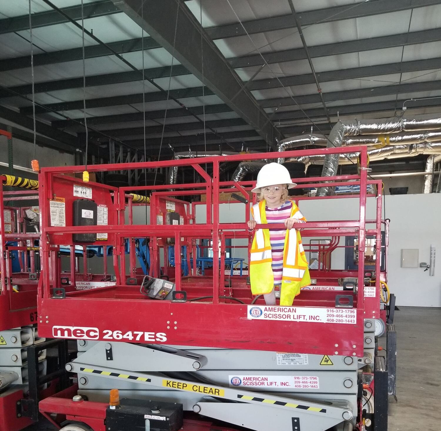 Child in a hard hat and safety vest standing on a red MEC 2647ES scissor lift platform