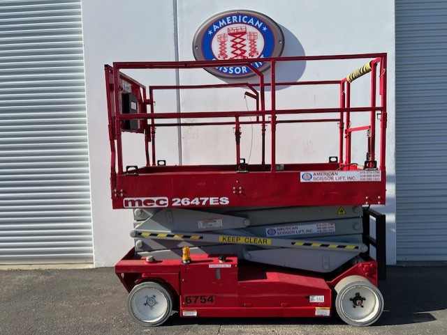 Used 2013 red MEC 2647ES electric scissor lift with its platform lowered in front of an American Scissor Lift building
