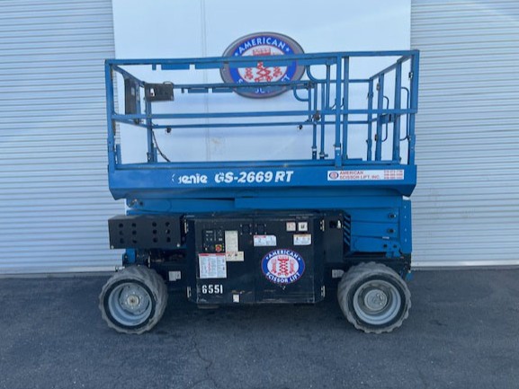 Used 2014 blue Genie GS-2669 RT rough terrain scissor lift with its platform lowered in front of an American Scissor Lift building