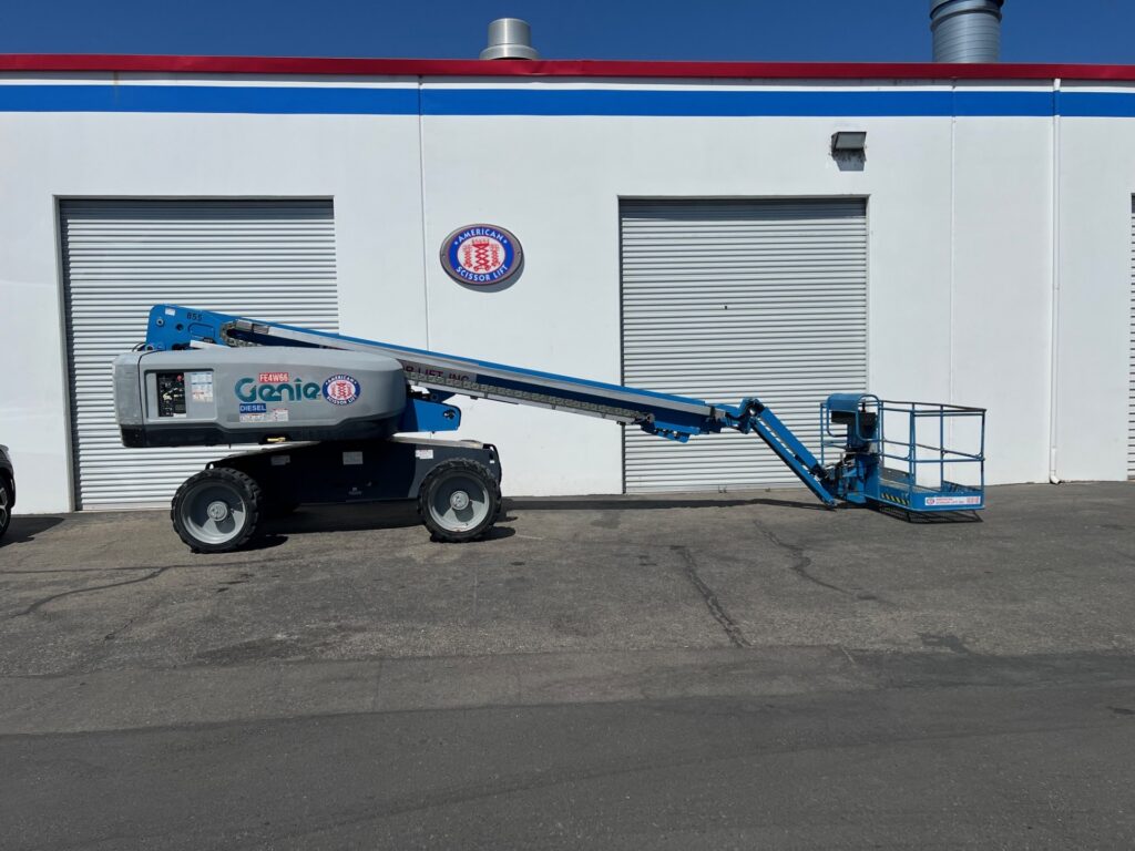 Used 2015 Genie S-65 telescopic boom lift with its boom lowered, parked in front of a warehouse, from the American Scissor Lift rental fleet