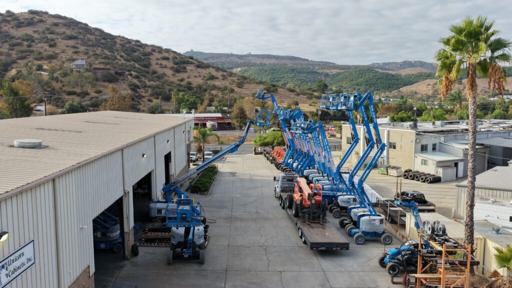 Aerial view of an American Scissor Lift rental yard with rows of blue Genie boom lifts raised beside the shop building and orange telehandlers on a flatbed trailer